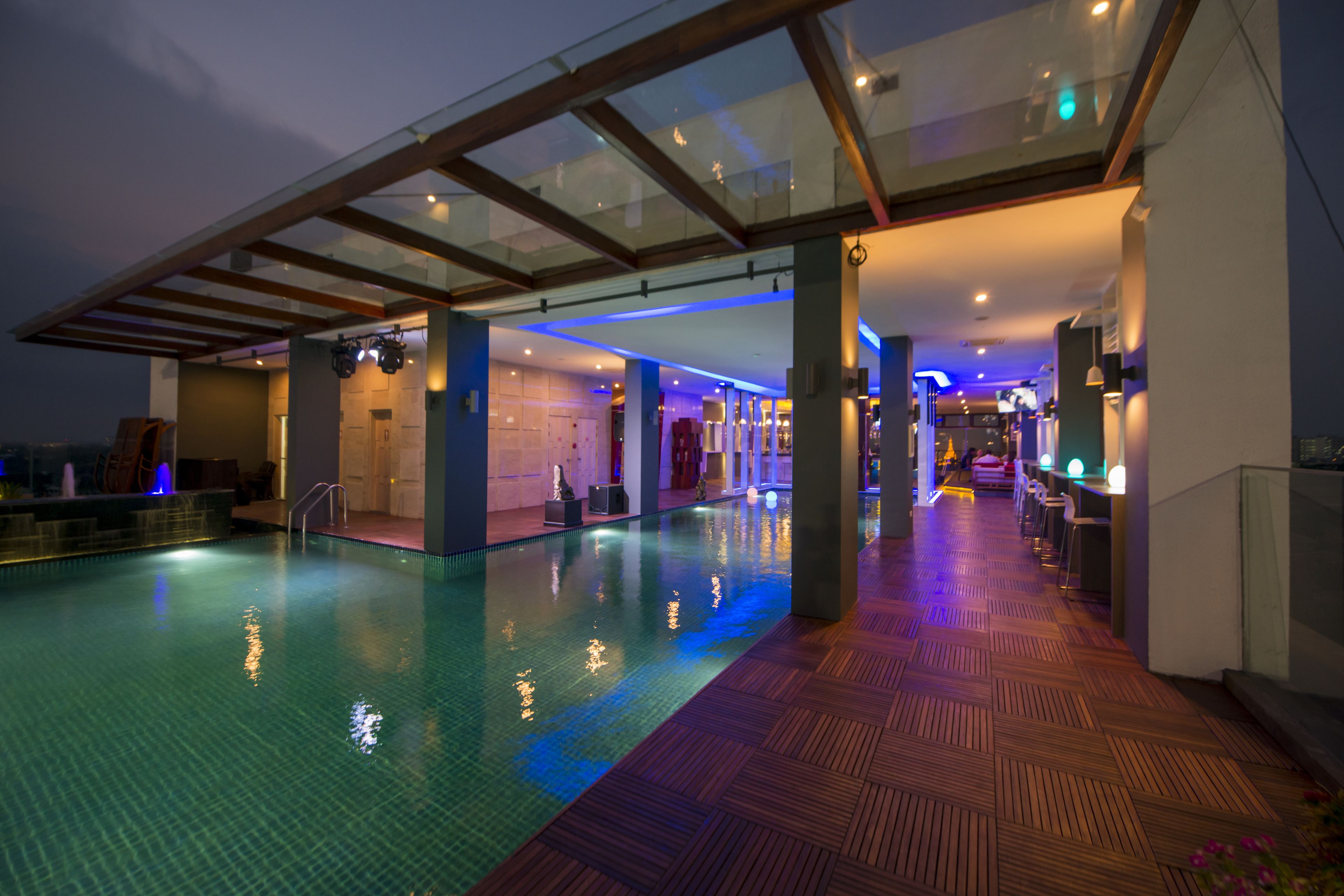 Una amplia piscina interior con solárium de madera, techo de cristal e iluminación moderna.