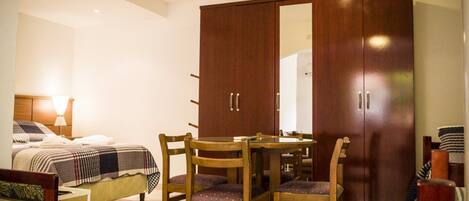 A bedroom with a bed, a wooden wardrobe, a dining table with chairs, and a bedside lamp.