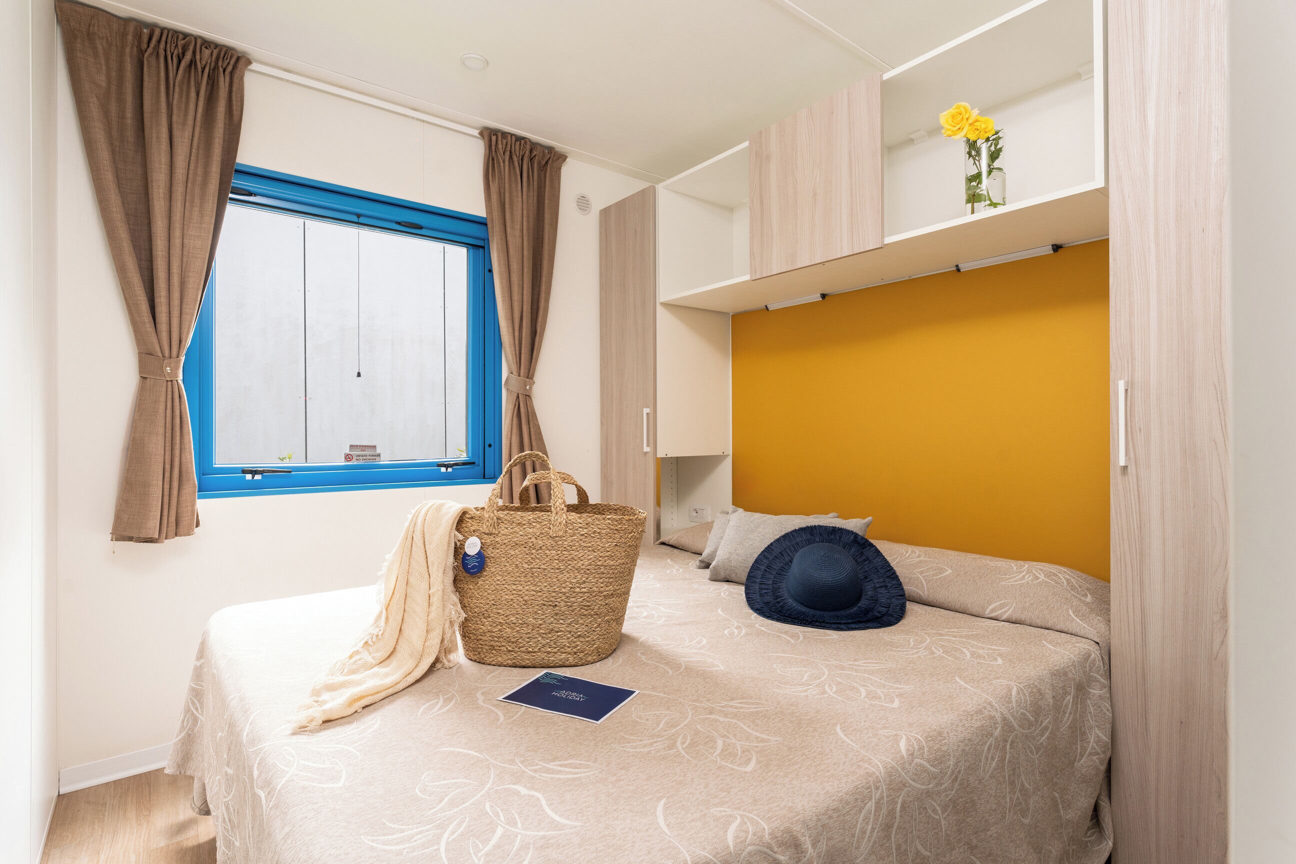 A bedroom with a bed, a window with curtains, a bedside table with a lamp, and a shelf with flowers.