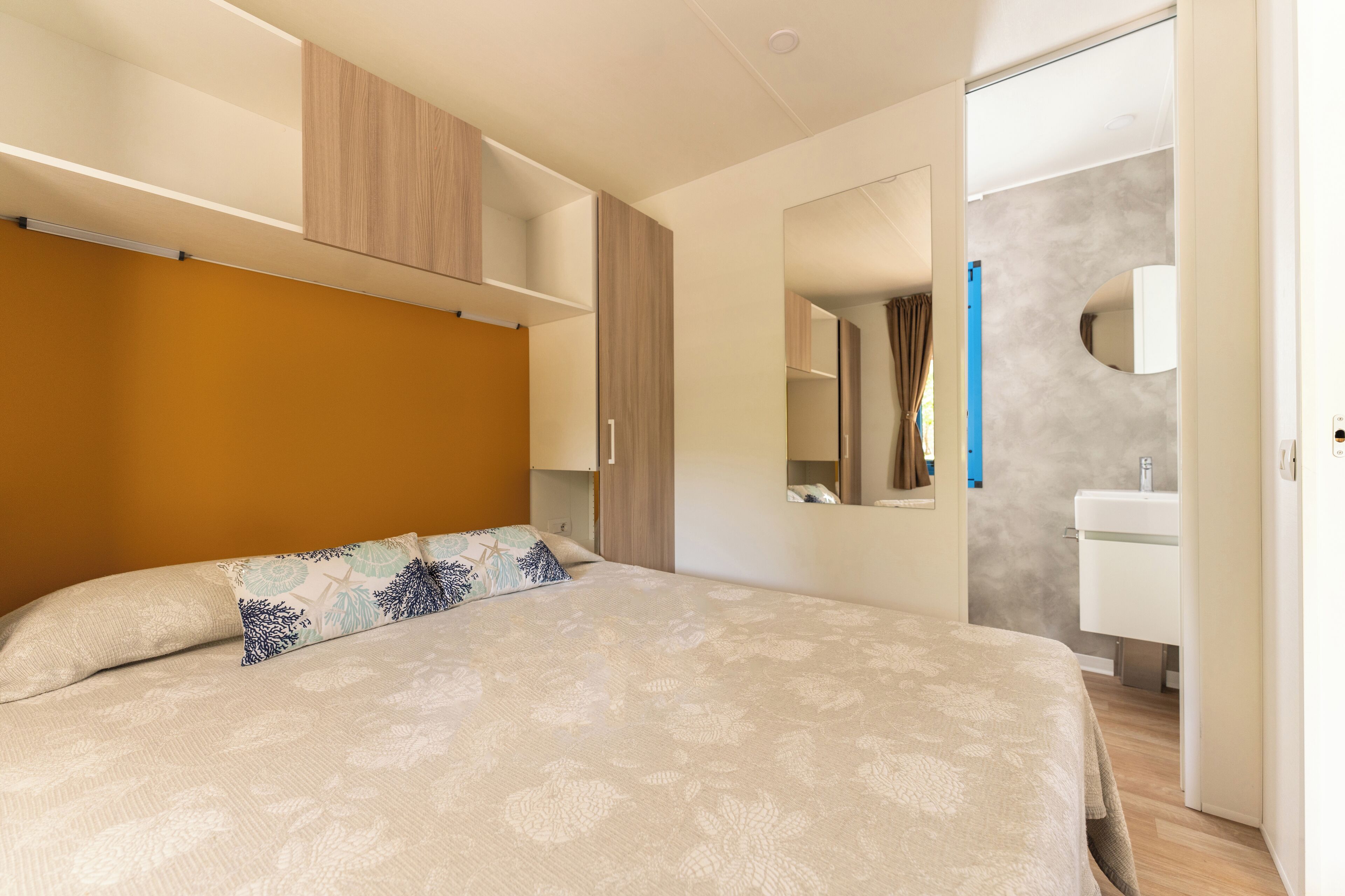 Une chambre à coucher avec un lit, une armoire, un miroir et une salle de bain visible à travers une porte ouverte.