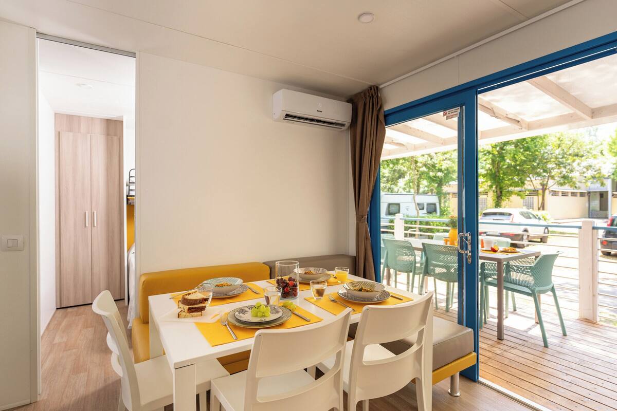 A dining area with a table set for four, a window with curtains, and an air conditioning unit on the wall.