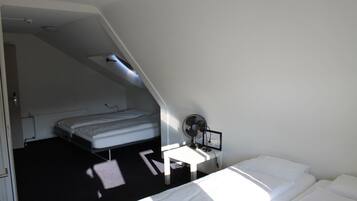 Une petite pièce faiblement éclairée, avec un lit simple, un bureau équipé d’un ordinateur et d’un ventilateur, et un plafond en pente.