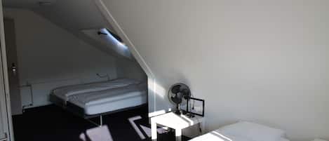 Een kleine, zwak verlichte kamer met een enkel bed, een bureau met een computer en ventilator, en een schuine plafond.