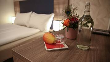 Una cama bien hecha con ropa blanca, una mesita de noche de madera con una botella de agua, un vaso de agua, un plato con un mango y un pequeño florero con flores.