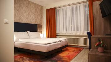 A modern hotel room with a bed, a desk, a chair, a TV, and a window with curtains.