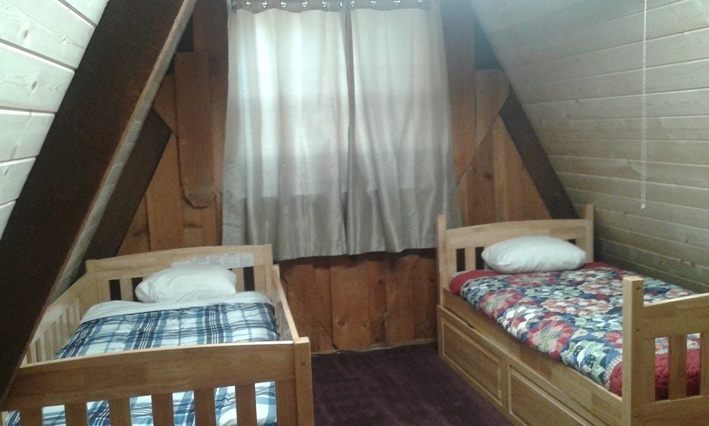 Un pequeño dormitorio en el ático con dos camas litera de madera, techo de madera y ventana con cortina translúcida.