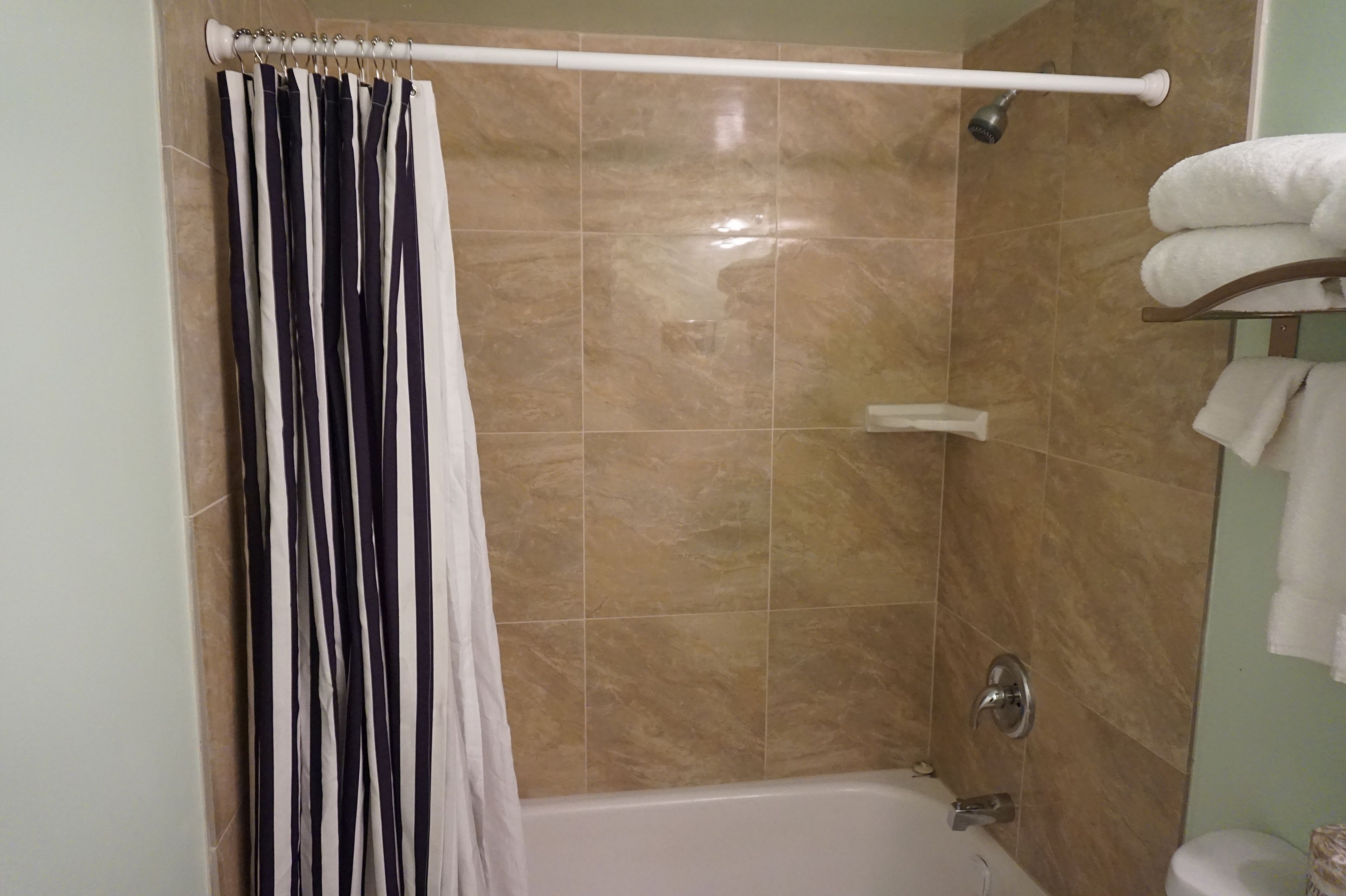 A shower area with a beige tiled wall, a white curtain, and a white towel rack with towels.
