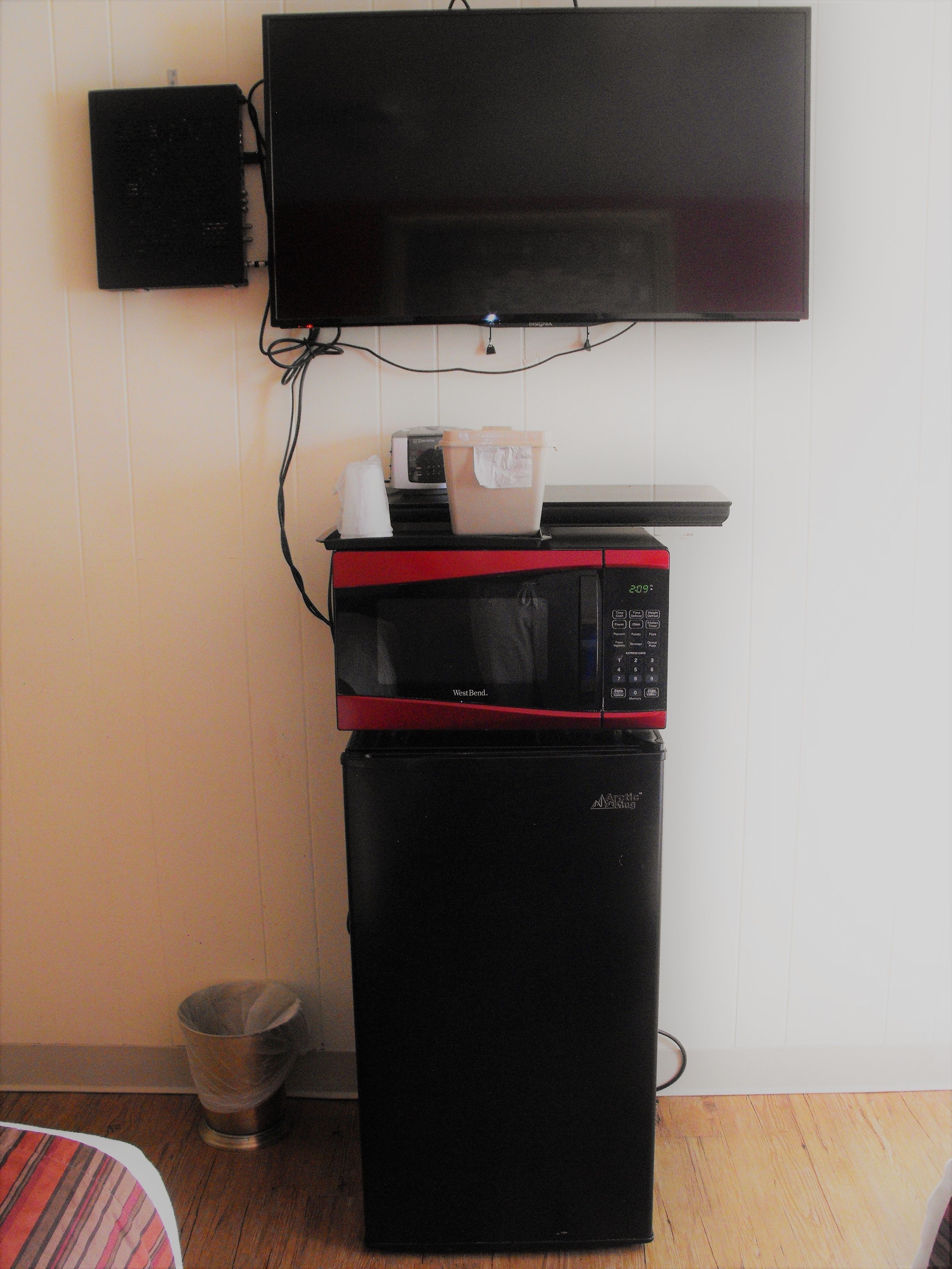 A compact kitchenette with a microwave, a small refrigerator, and a television mounted on the wall.