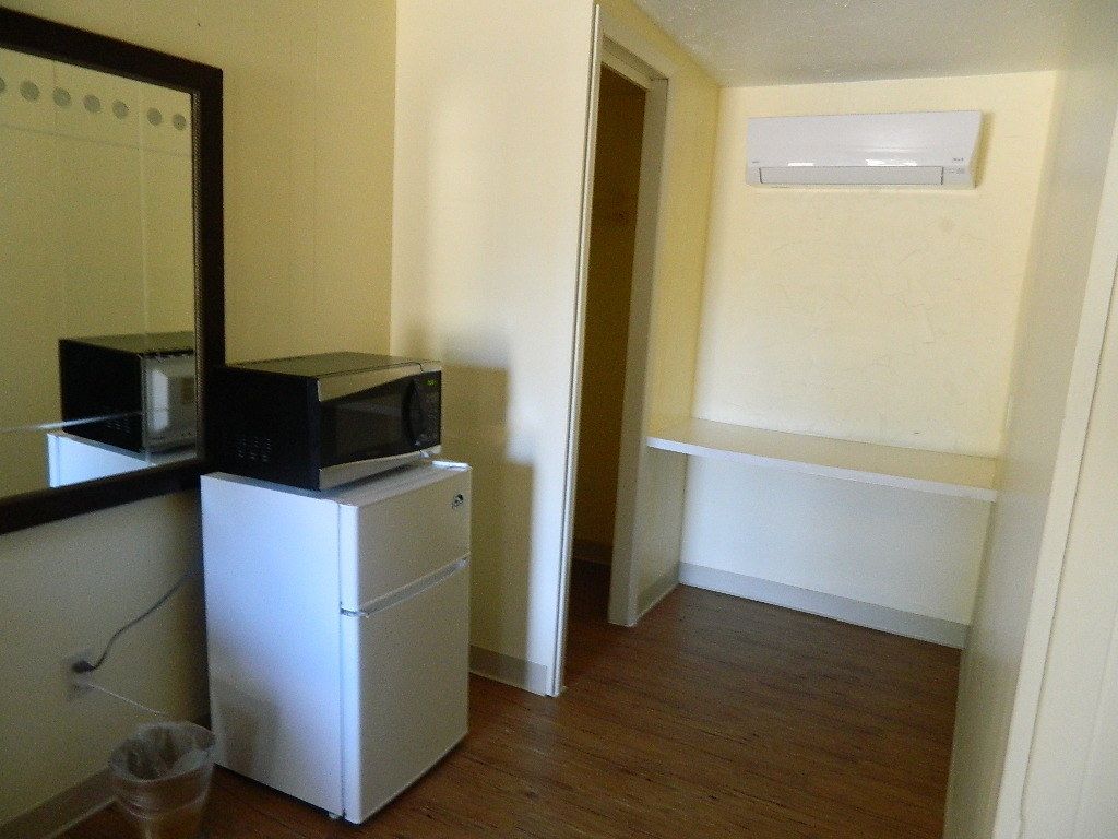 Una cocina compacta con microondas y refrigerador, piso de madera y pared blanca con una ventana pequeña.