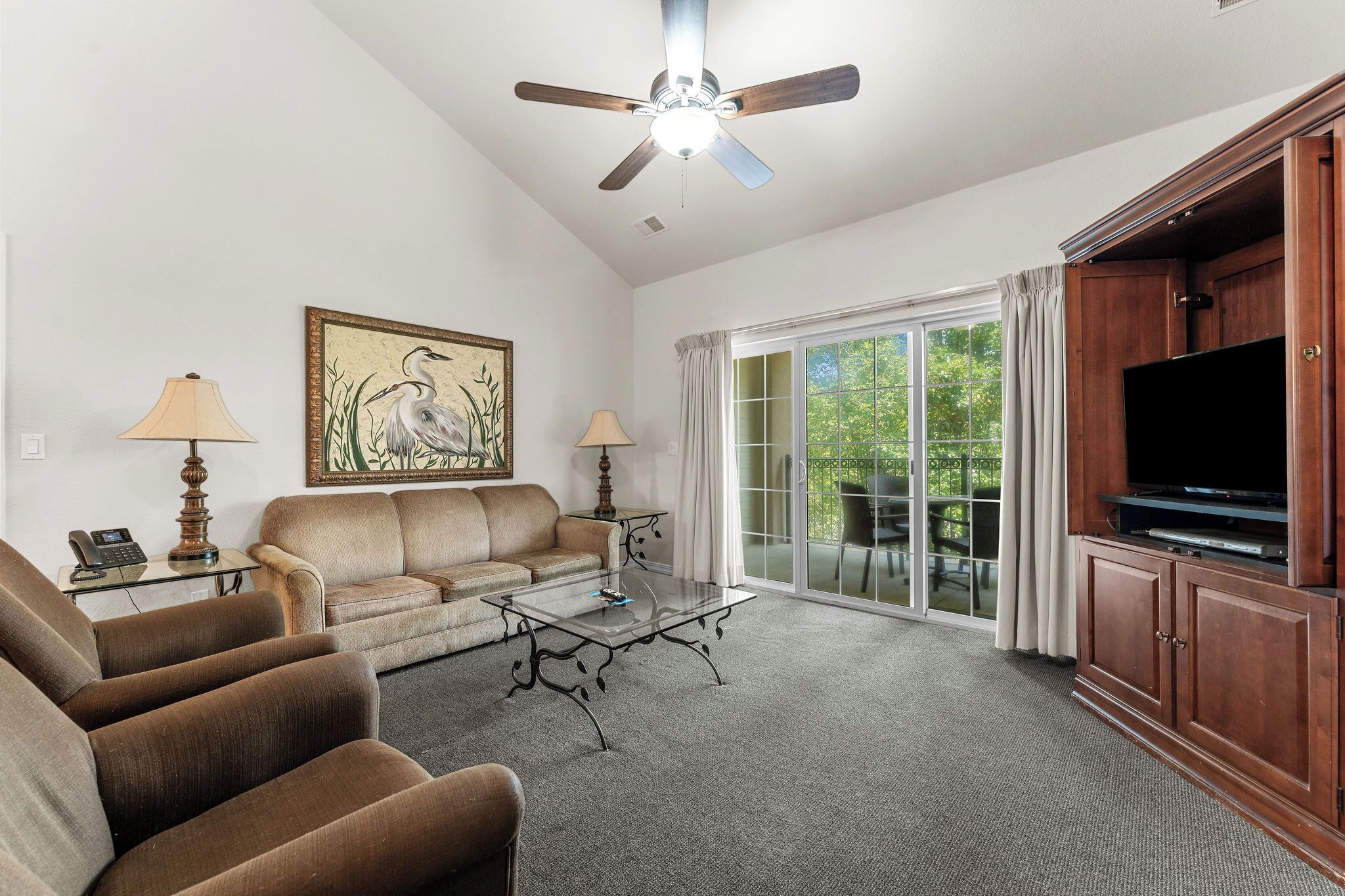 A living room with a sofa, armchairs, a coffee table, a ceiling fan, a painting, a television, and a sliding glass door.