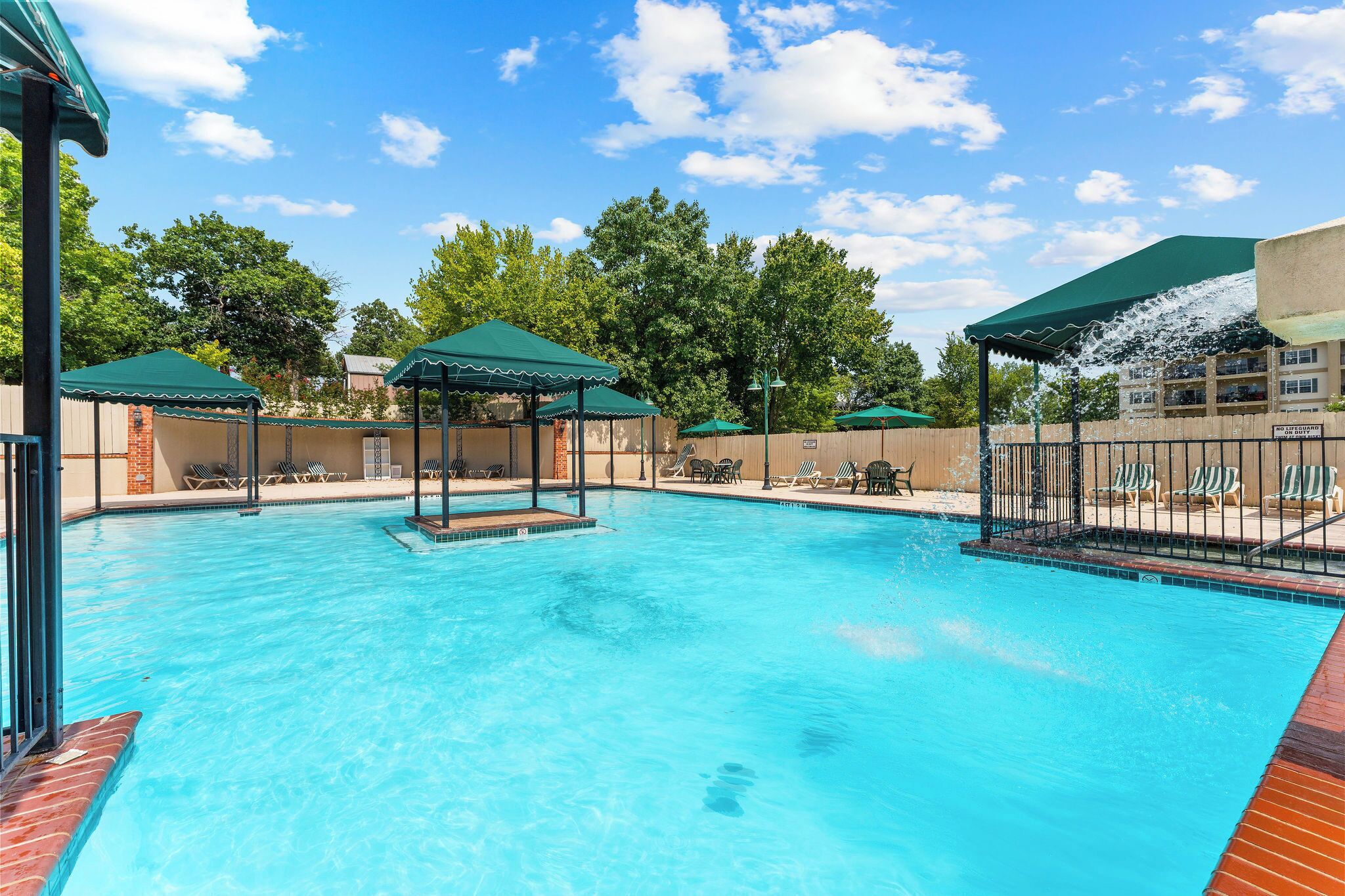 A swimming pool with lounge chairs and umbrellas, surrounded by a fence and trees.