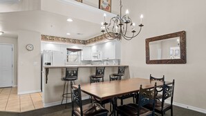 Un comedor con una mesa y sillas de madera, una araña y una cocina al fondo.