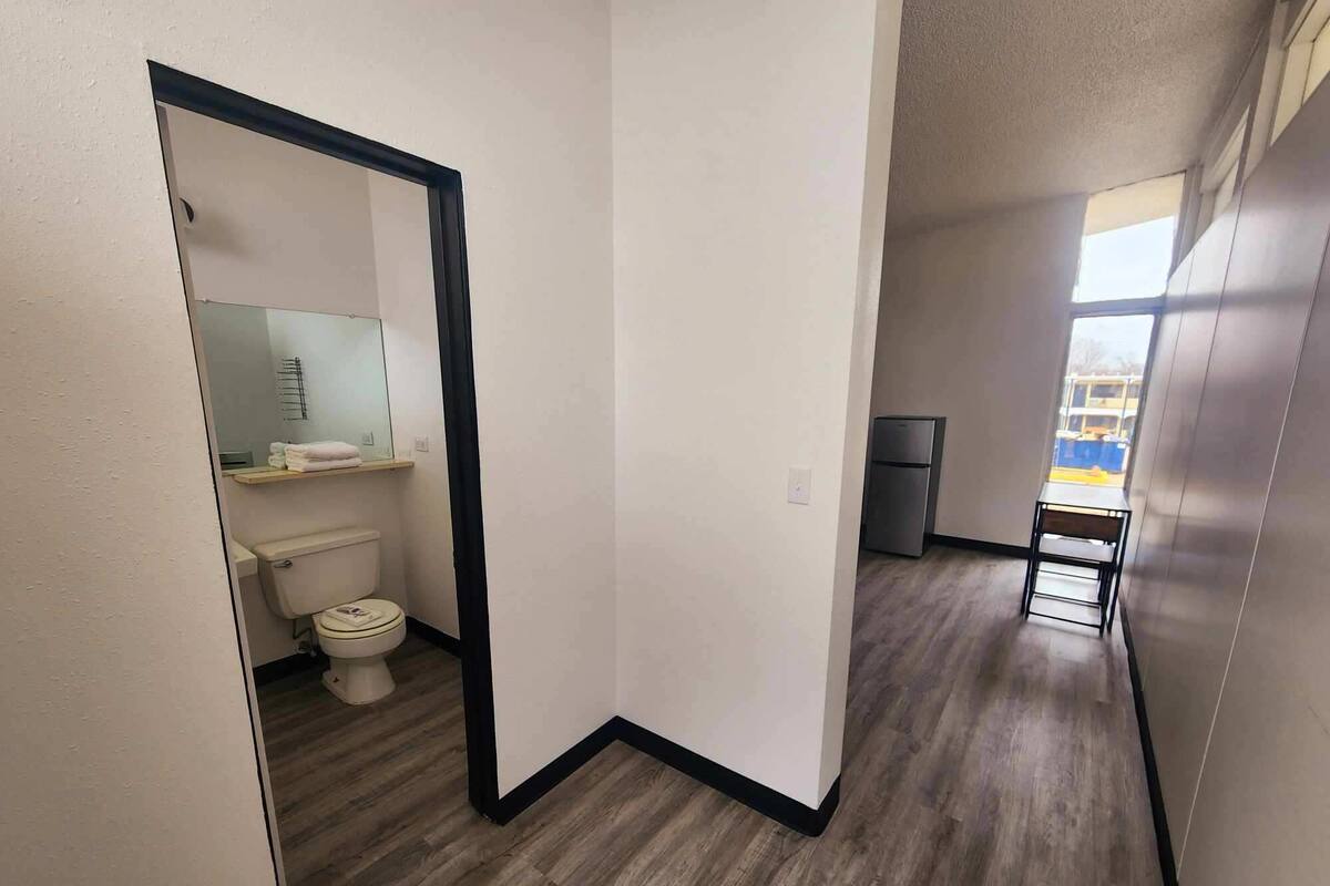 A narrow hallway with a wooden floor, leading to a bathroom with a toilet and a mirror.