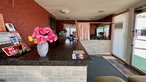 Zona de recepción de hotel con mostrador de mármol, cartel de 'No Fumar', un jarrón con flores rosas y un cartel que indica 'Llamar para Asistencia'.