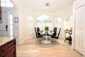 A dining area with a round table, four chairs, and a chandelier. There are windows with blinds, a door, and a small plant on a stand. - Crystal Cove by CozyKey Vacation Rentals (Kissimmee)