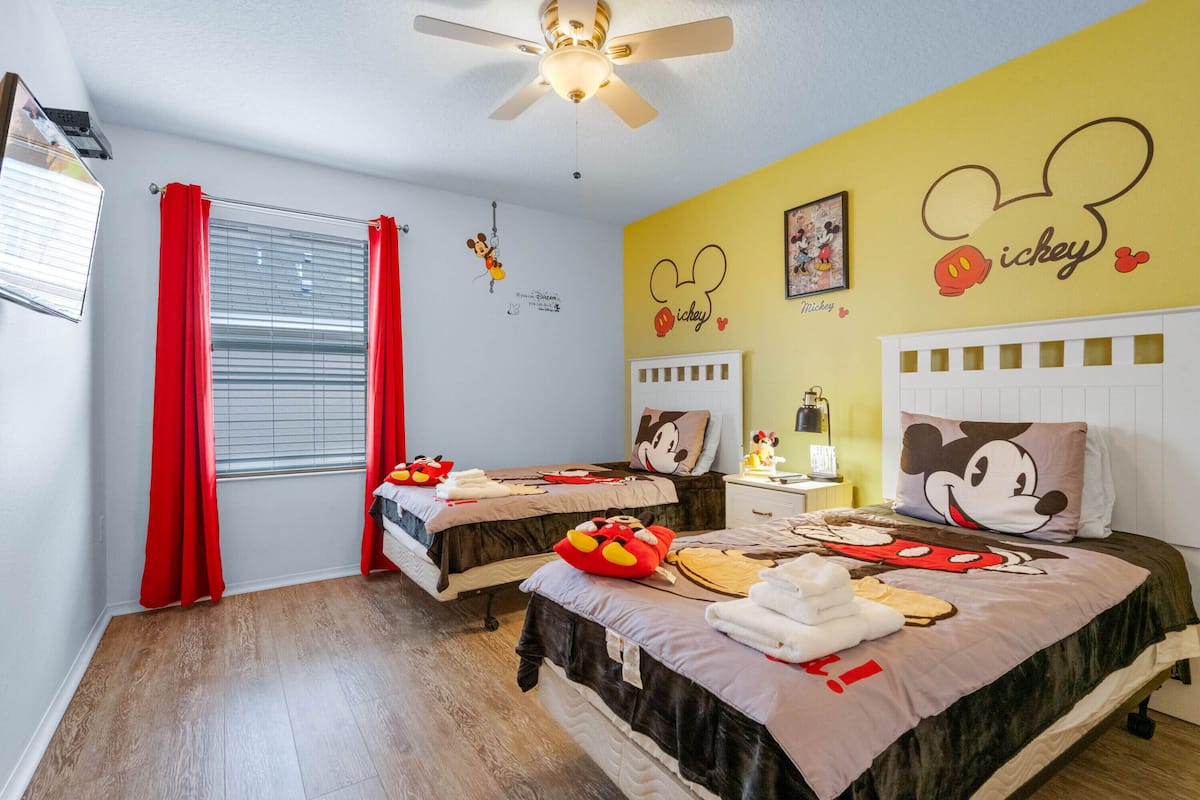 Une chambre d’hôtel avec deux lits, des rideaux rouges, des décorations à l’effigie de Mickey Mouse et un ventilateur de plafond.