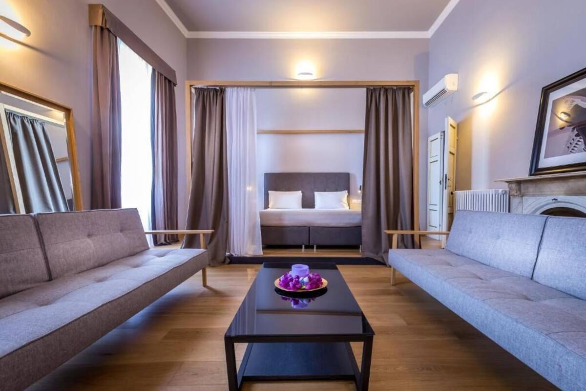A modern hotel room with a large bed, two sofas, a coffee table, and a fireplace.