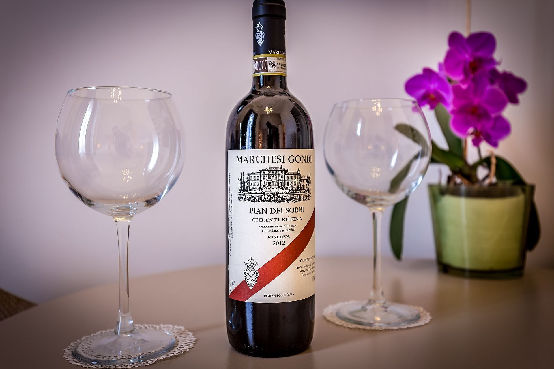 A bottle of wine with a label, two empty wine glasses, and a potted orchid.