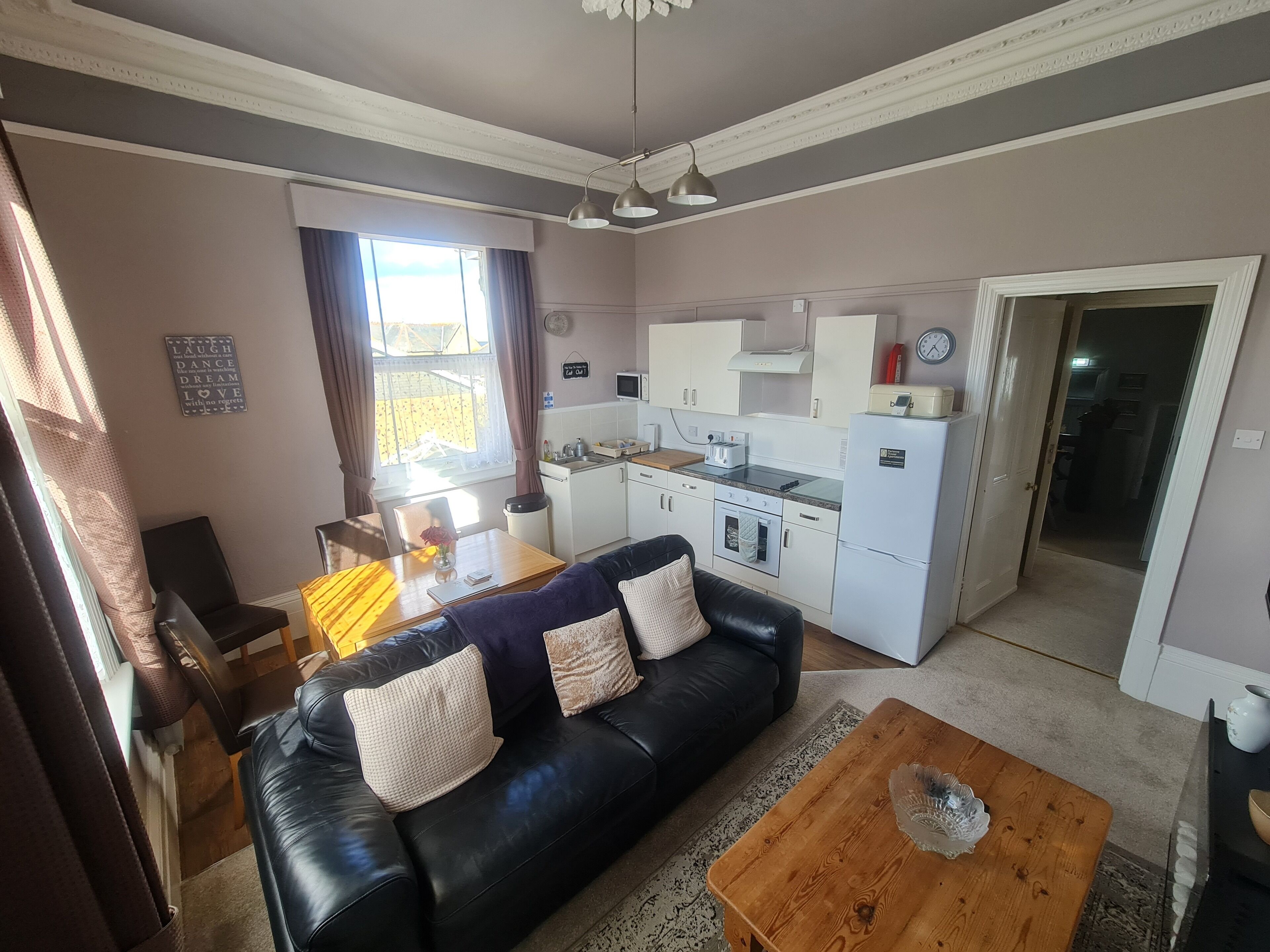 Un espacio habitable compacto con un sofá de cuero negro, una mesa de centro de madera, una mesa de comedor con sillas, una kitchenette y un ventanal con cortinas.