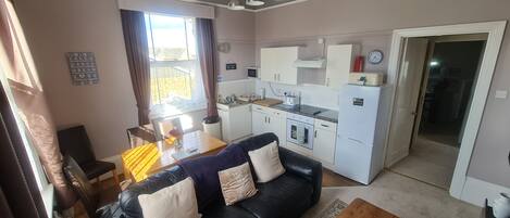 Un espacio habitable compacto con un sofá de cuero negro, una mesa de centro de madera, una mesa de comedor con sillas, una kitchenette y un ventanal con cortinas.