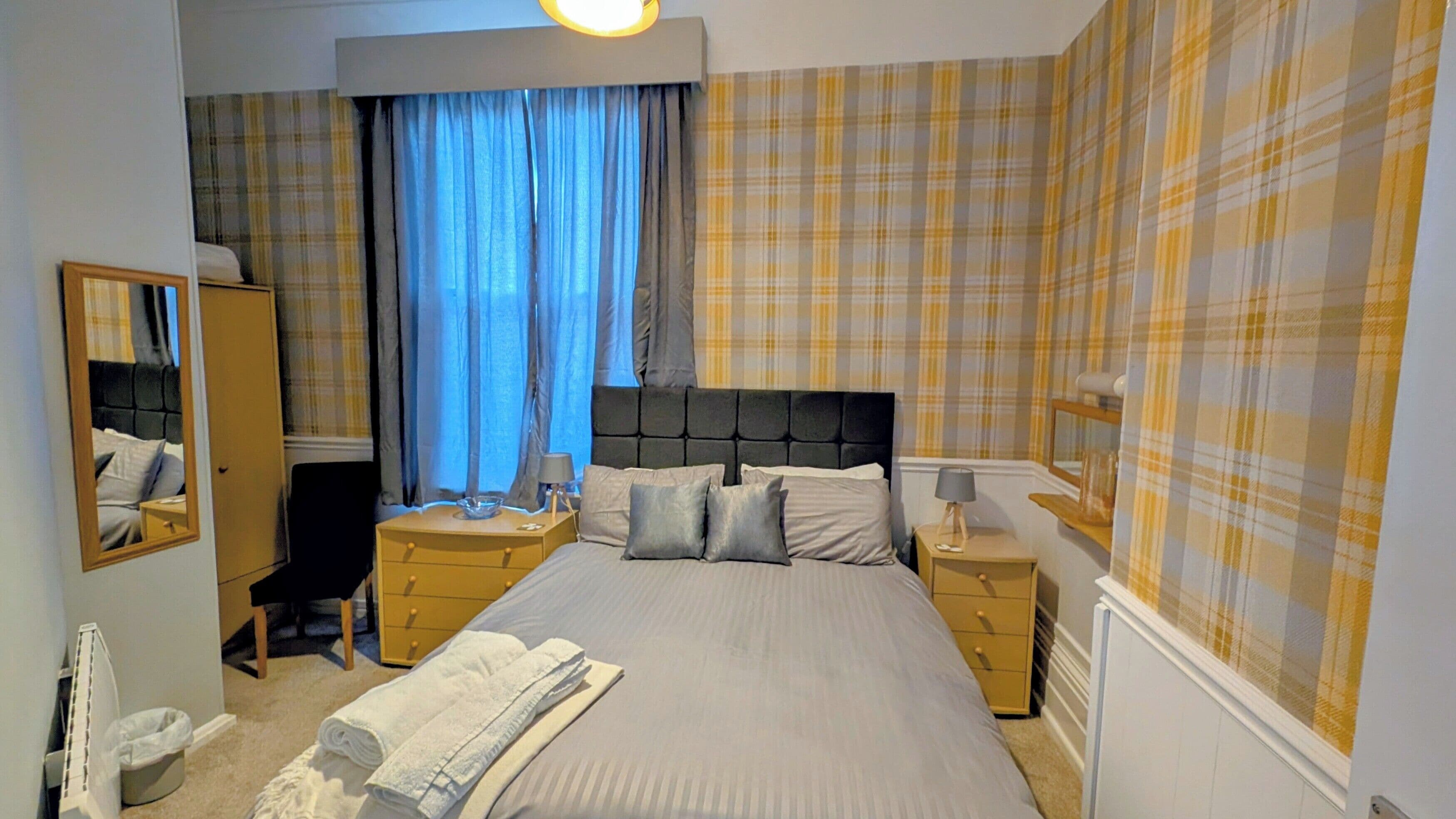A bedroom with a bed, two bedside tables, a mirror, a wardrobe, and a window with curtains.