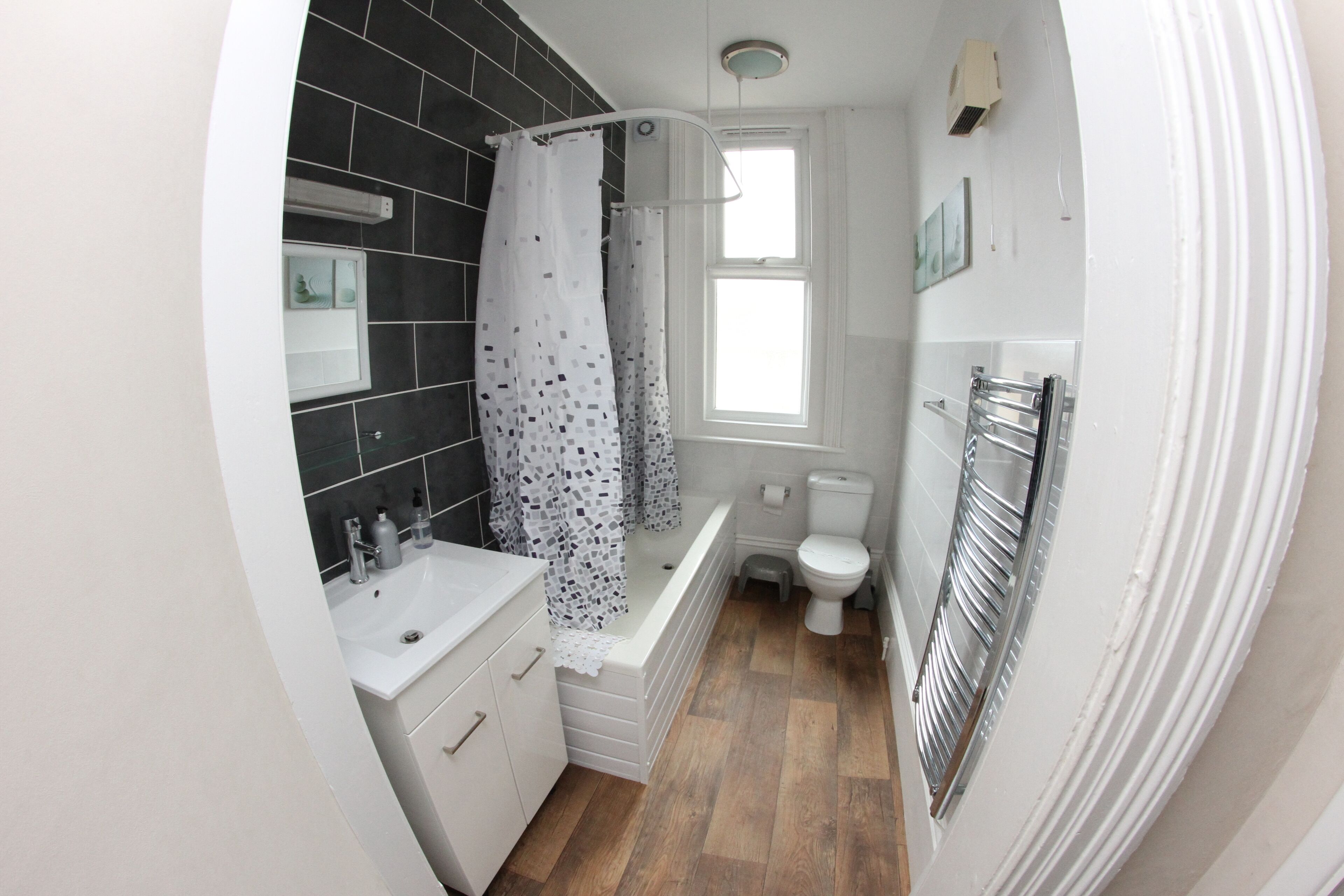 A modern bathroom with a white sink, black tiled walls, a shower with a patterned curtain, a toilet, and a window.