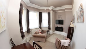 A living room with a sofa, a dining table, and a TV. - Parterre Holiday Apartments (Sandown)