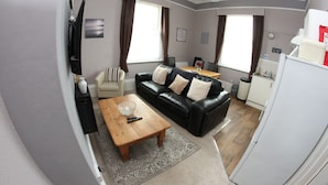 A living room with a black leather sofa, a wooden coffee table, and a television mounted on the wall. - Parterre Holiday Apartments (Sandown)
