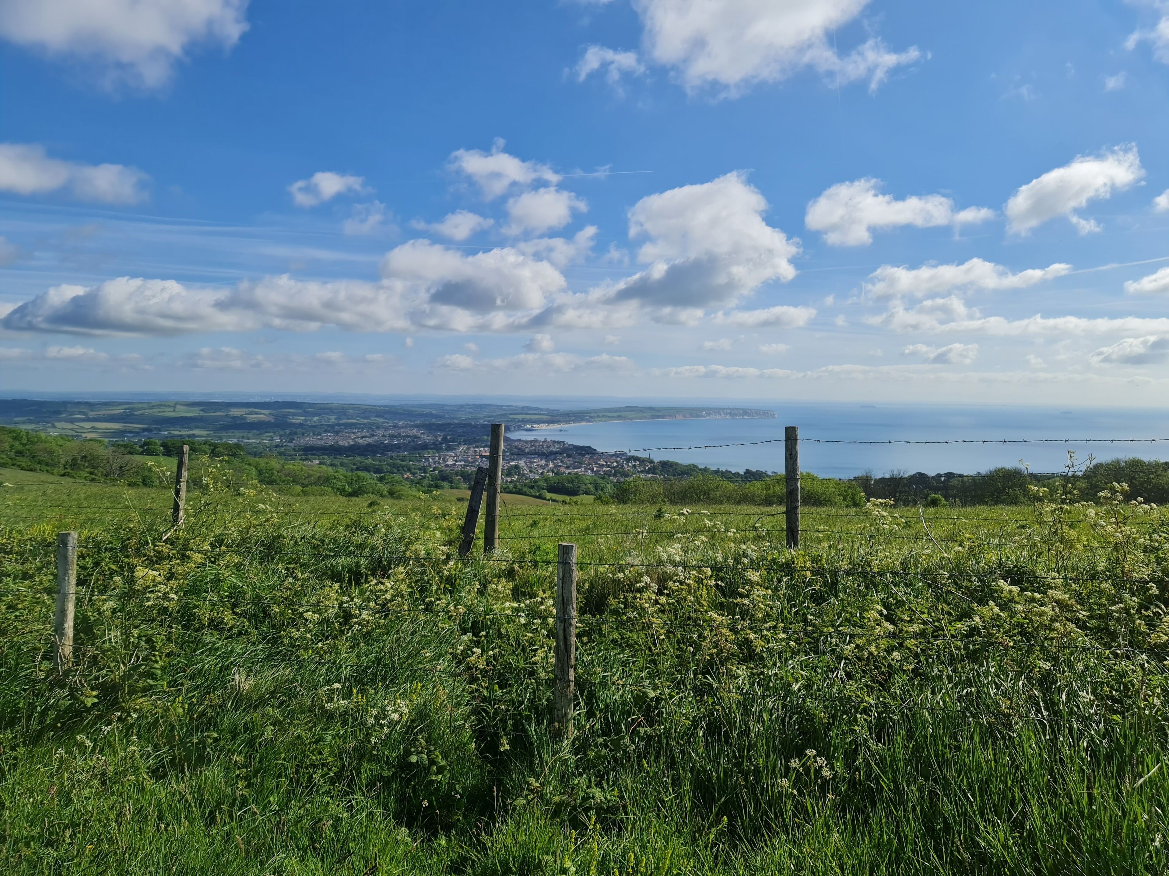 Ein grasbewachsener Hügel mit Holzzaun und weiter Aussicht auf eine Küstenstadt und das Meer.
