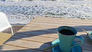 A wooden deck with a table and chairs, a cup of coffee, and a view of the ocean. - Parterre Holiday Apartments (Sandown)