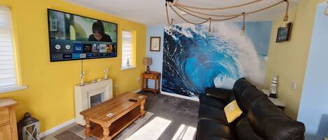 A living room with a large TV mounted on the wall, a fireplace, a black leather sofa, and a mural of a wave on the wall.