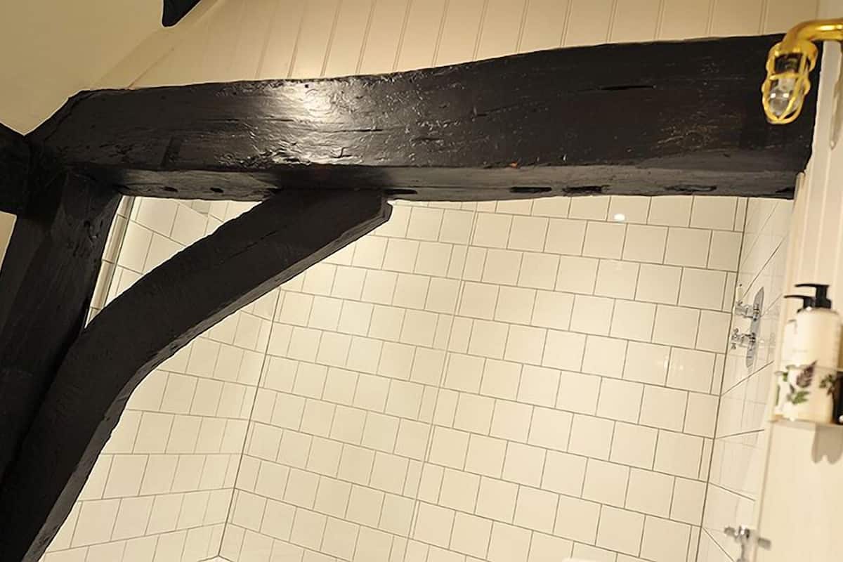 Un baño con paredes de azulejos blancos, estructura de vigas de madera negra y una zona de ducha con cabezal dorado.