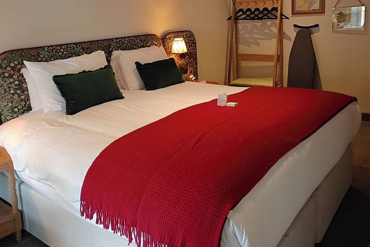 Una cama bien hecha con sábanas blancas y una manta roja, una mesita de noche con lámpara, y un perchero en la pared.
