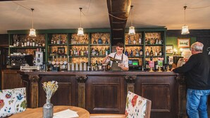 Un bar con paneles de madera, estantes llenos de botellas y una barra donde un barman prepara una bebida. Hay dos clientes sentados en una mesa con un jarrón de flores.