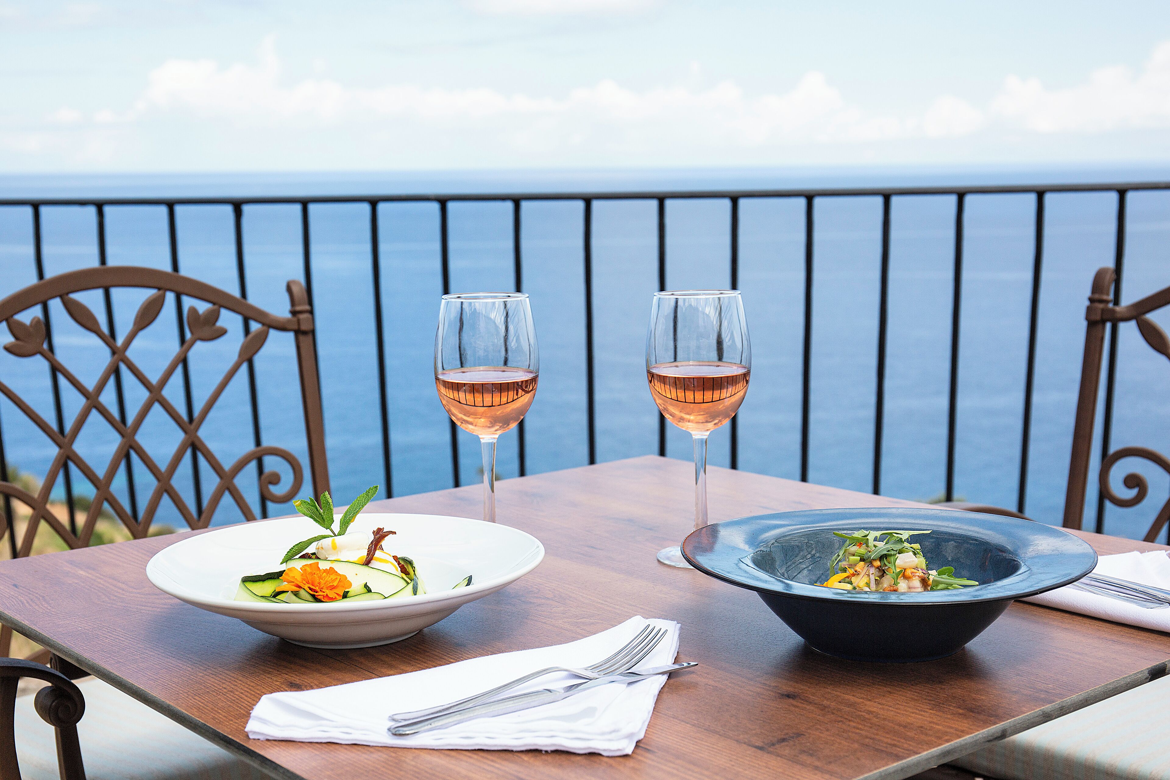 A table with two bowls of food and two glasses of wine, set on a balcony with a view of the sea.