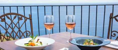 A table with two bowls of food and two glasses of wine, set on a balcony with a view of the sea.