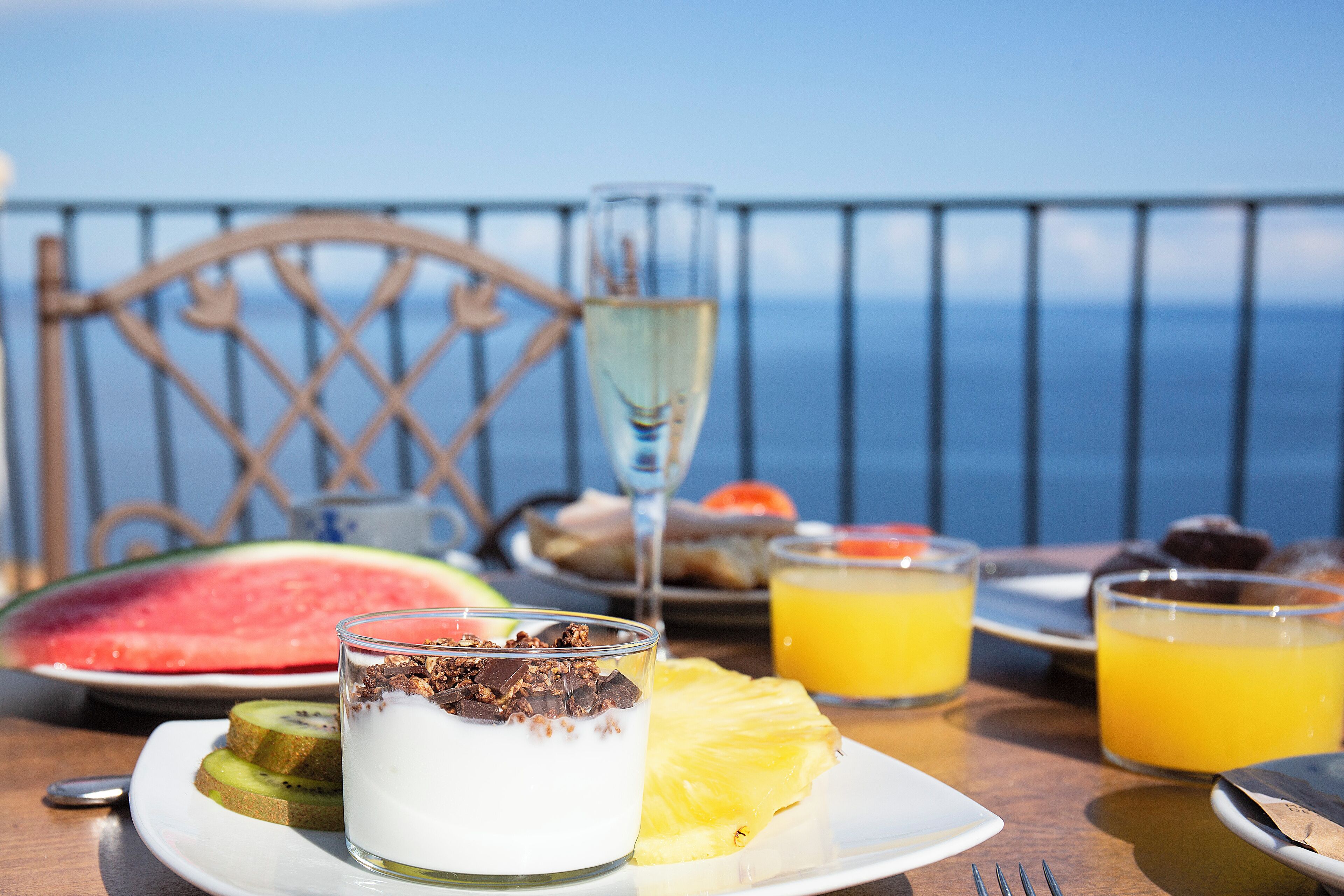A breakfast table with a plate of yogurt, a slice of watermelon, a glass of orange juice, and a glass of champagne.