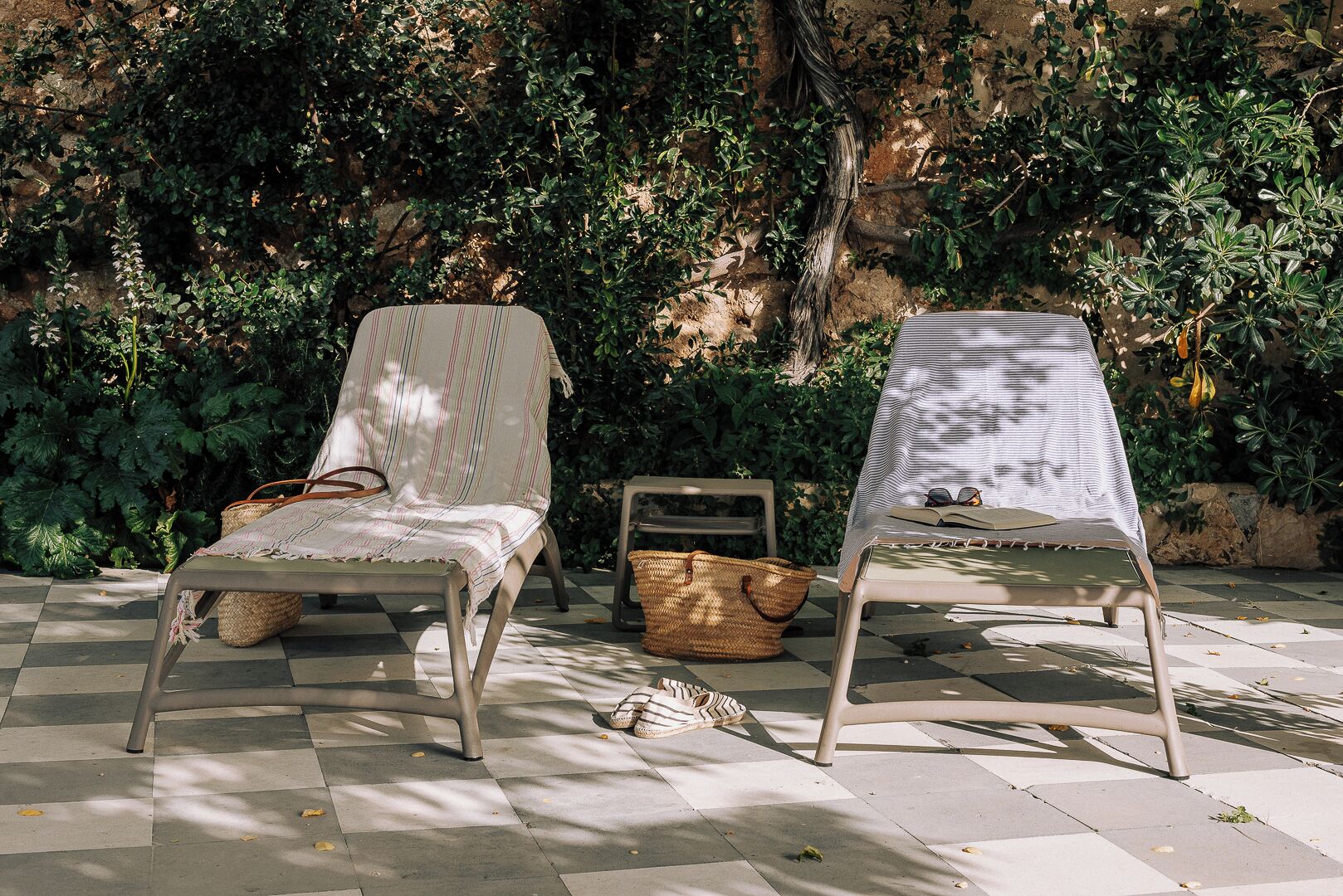 Deux fauteuils de jardin avec des coussins à rayures, un panier en osier et un livre posé sur une table, le tout installé sur un sol extérieur à carreaux, avec de la verdure en arrière-plan.