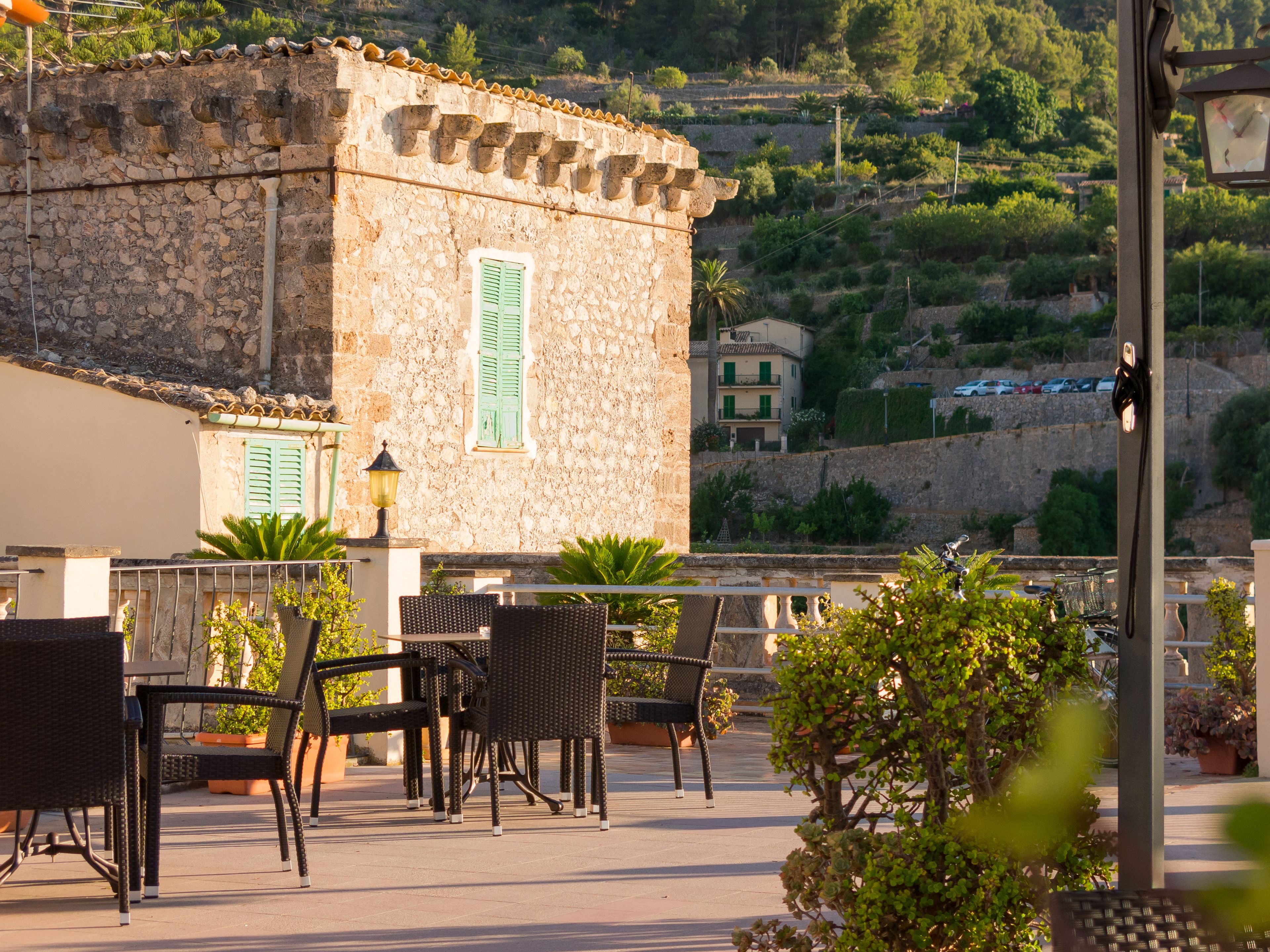 A terrace with wicker chairs and potted plants, overlooking a stone building with green shutters and a view of a hillside with houses.