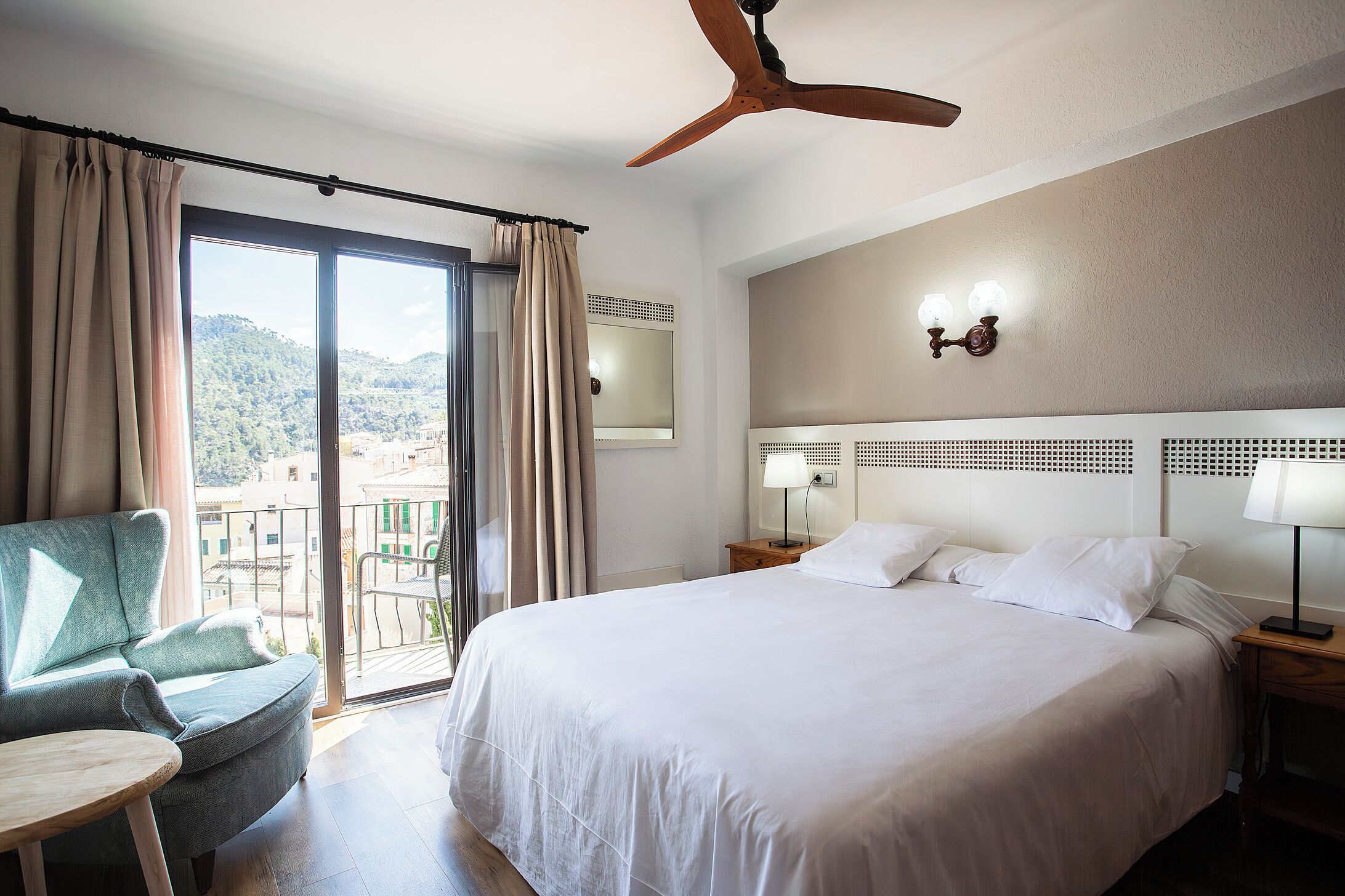 A hotel room with a bed, a chair, a table, a ceiling fan, and a balcony with a view.