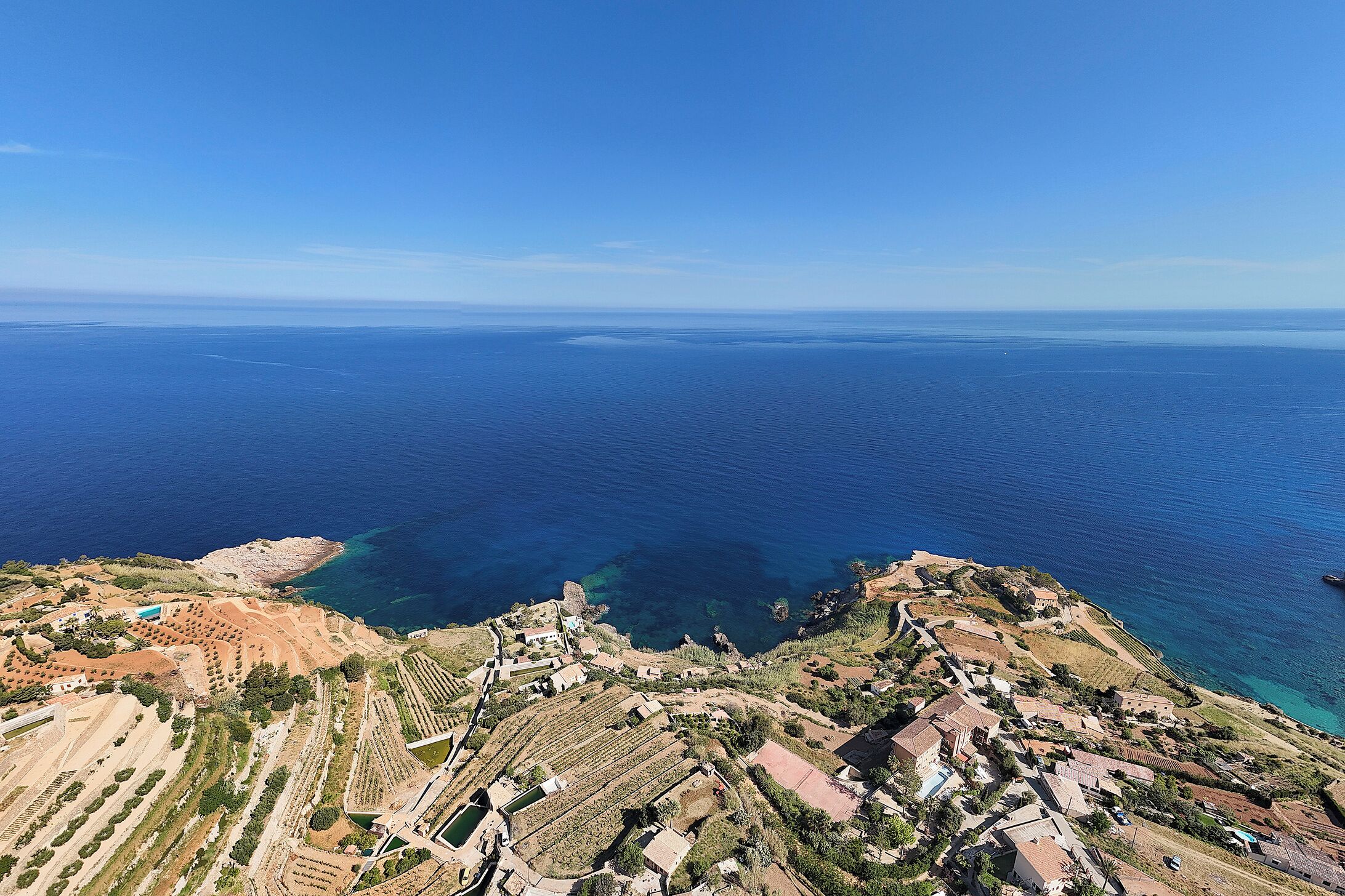 An aerial view of a coastal area with terraced vineyards, buildings, and a clear view of the sea.