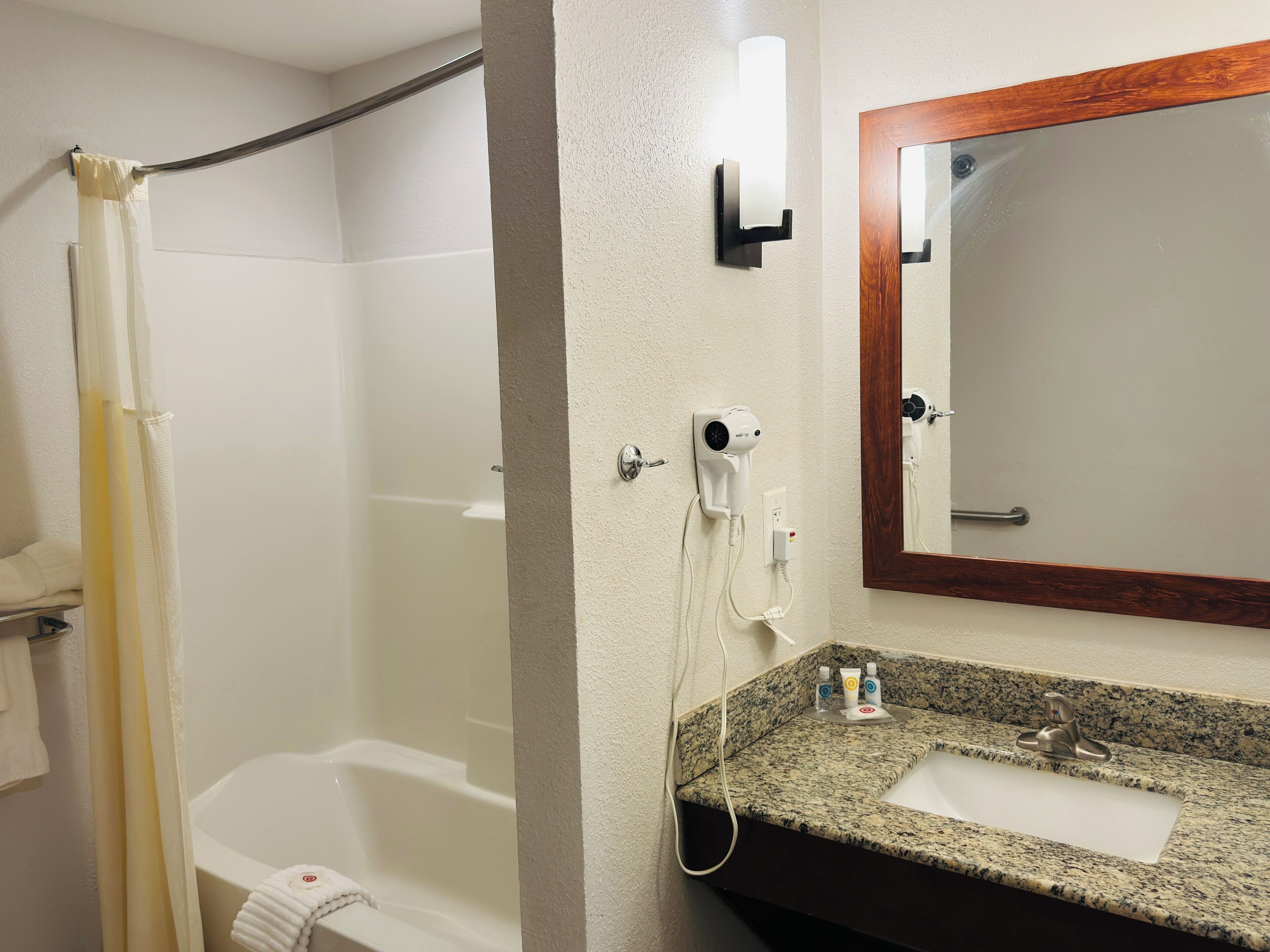 A bathroom with a shower, a sink with a granite countertop, a mirror with a wooden frame, and a hairdryer mounted on the wall.