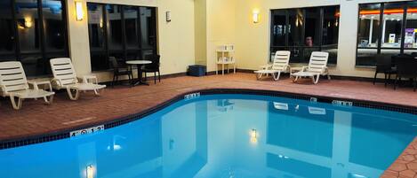 A swimming pool with lounge chairs and tables, surrounded by windows and a tiled floor.