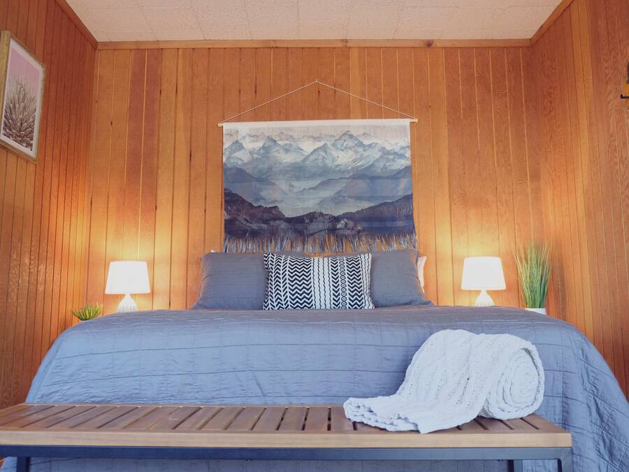A wooden-paneled room with a bed, two bedside lamps, a painting of mountains, and a small table with a blanket.
