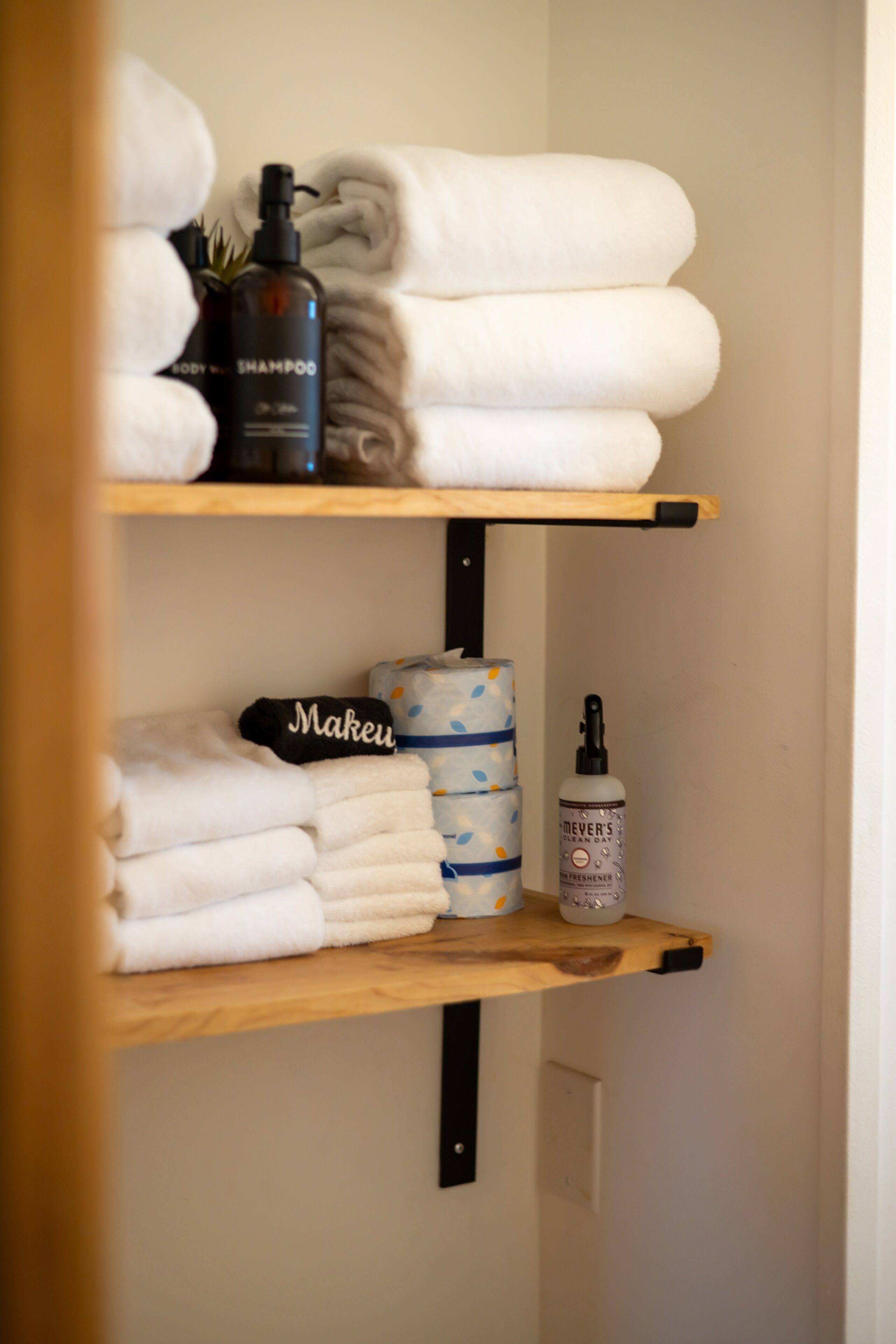 A shelf with neatly folded towels, a bottle of shampoo, a bottle of body spray, and a roll of toilet paper.