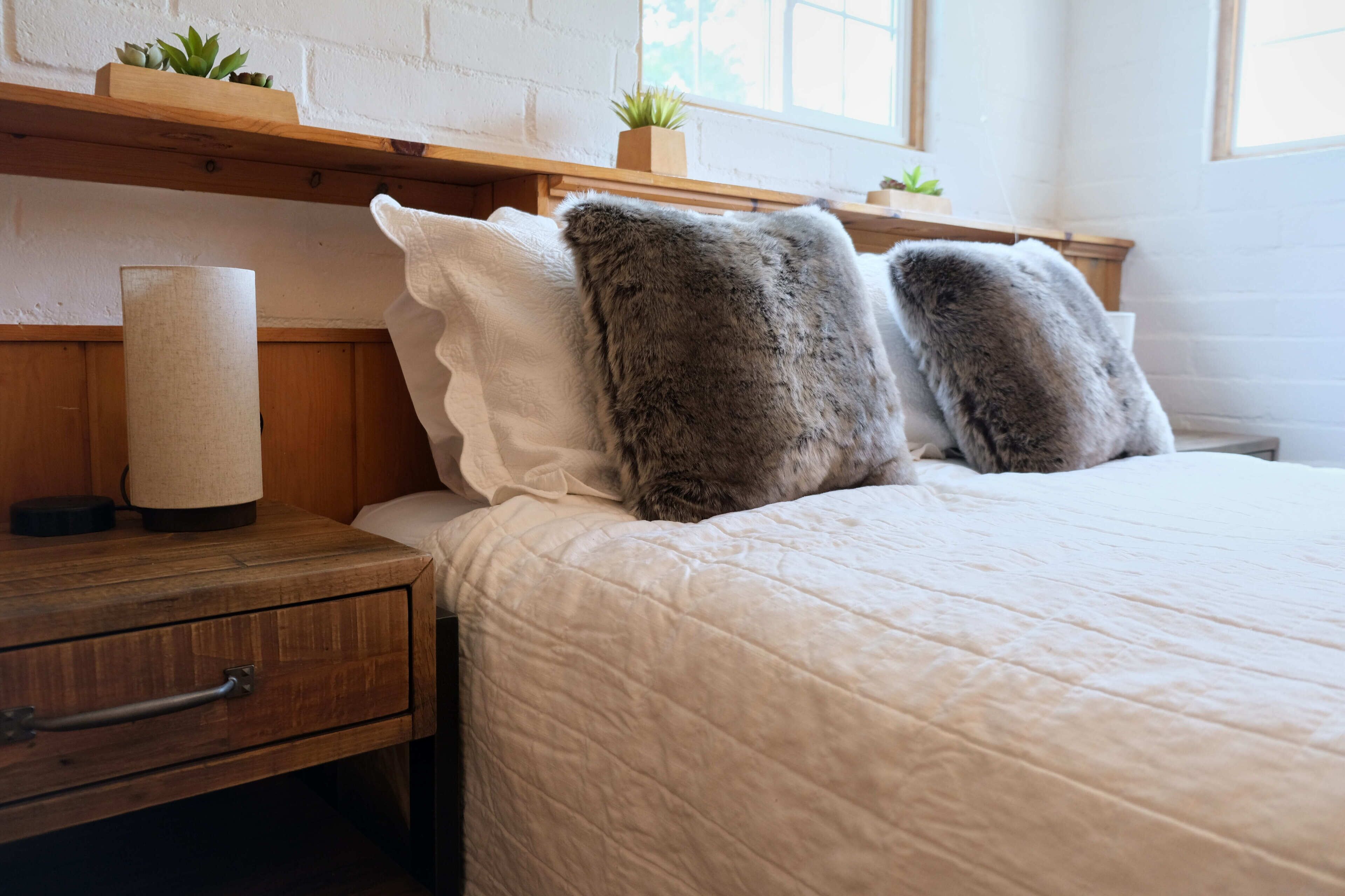 A neatly made bed with fluffy pillows, a wooden nightstand with a lamp, and a small plant on the shelf above.