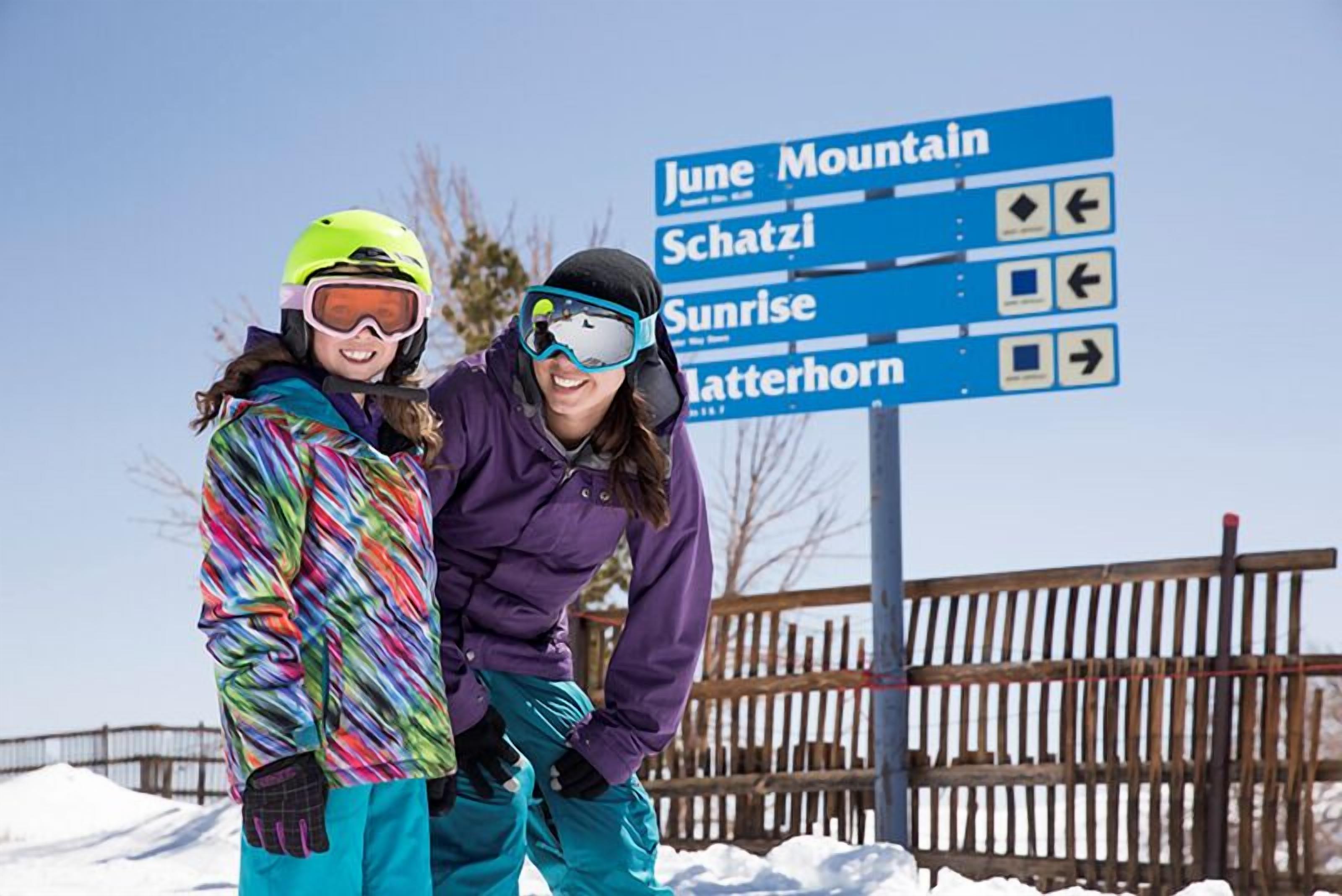 Deux personnes, vêtues de vestes d’hiver colorées, se tiennent devant un panneau indicateur indiquant des destinations telles que June Mountain et Sunrise.