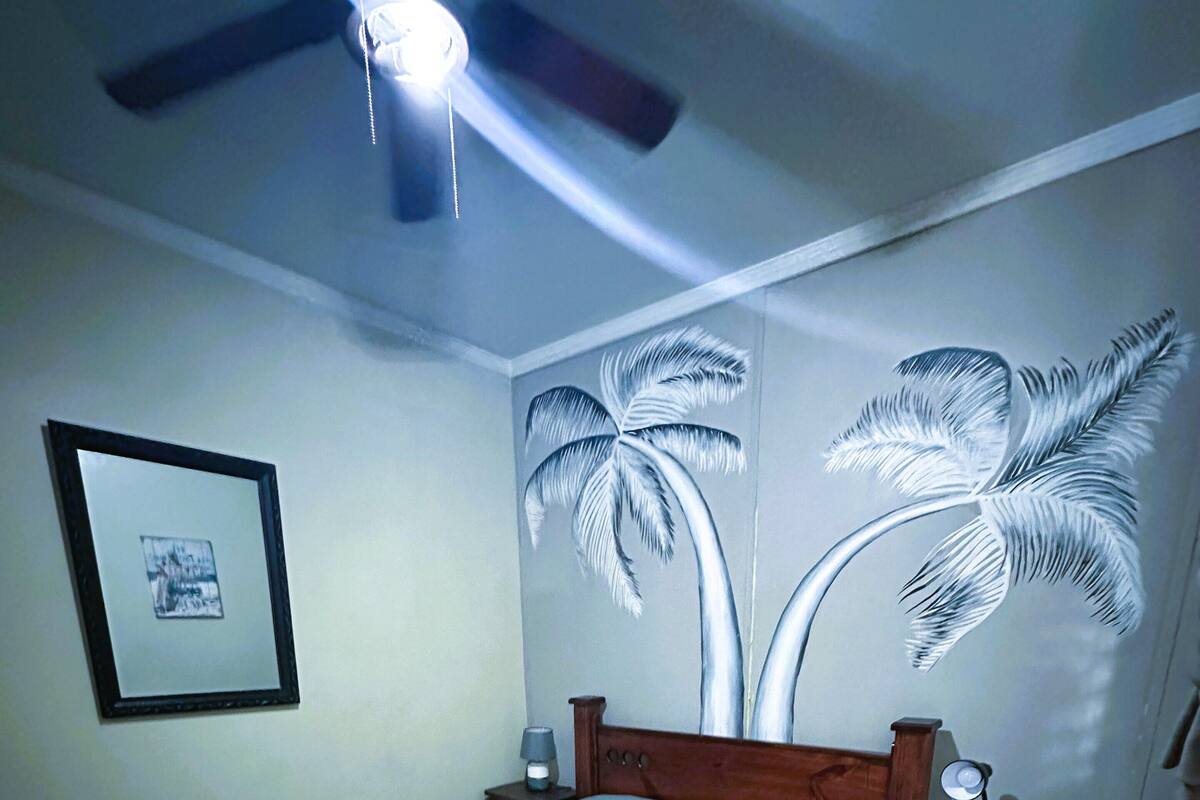A bedroom with a bed, a nightstand, a framed picture, and a wall mural of palm trees.