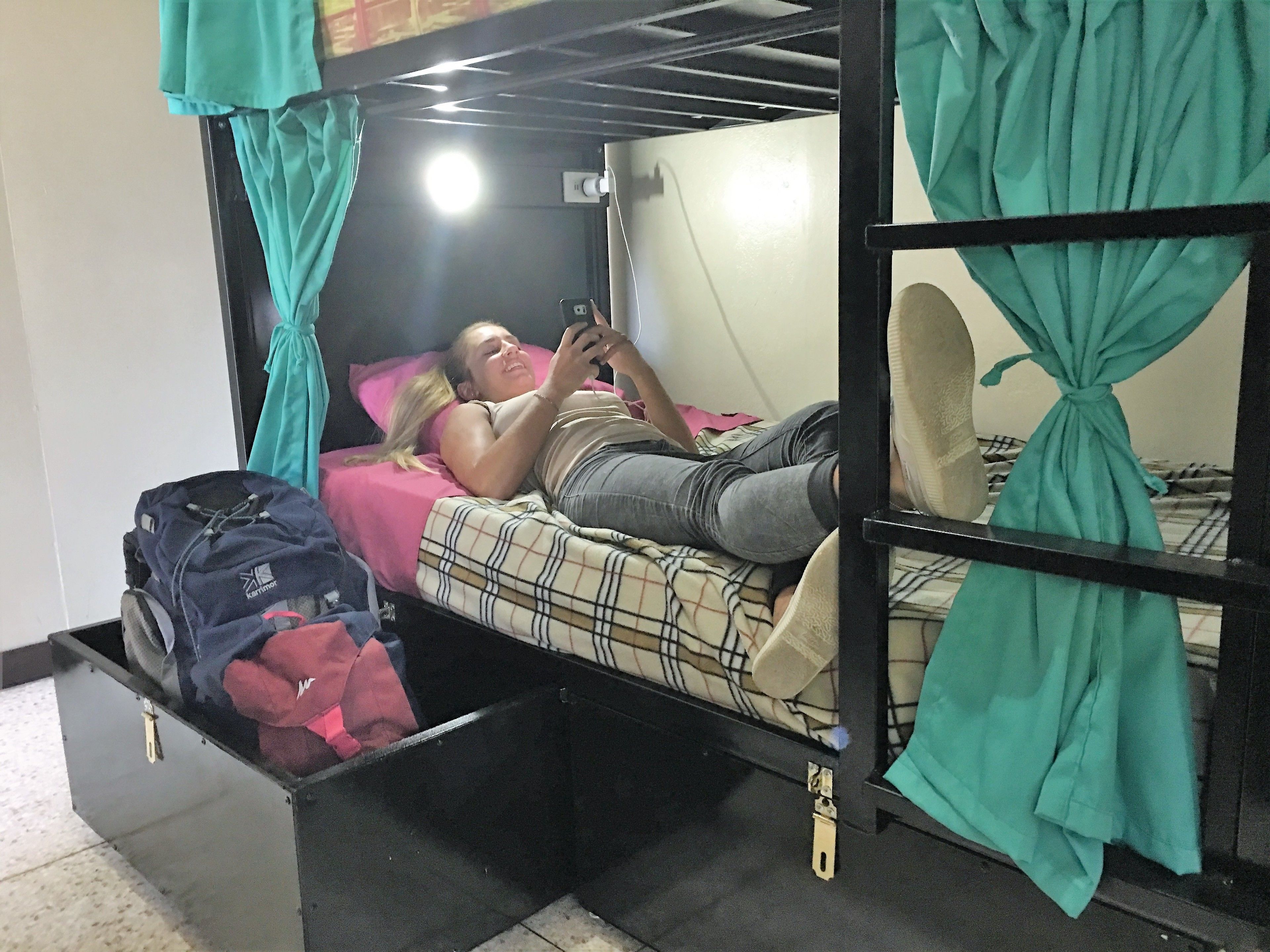 Una cama individual con cortinas color turquesa, una persona acostada en la cama usando un teléfono móvil, y una maleta en el suelo.