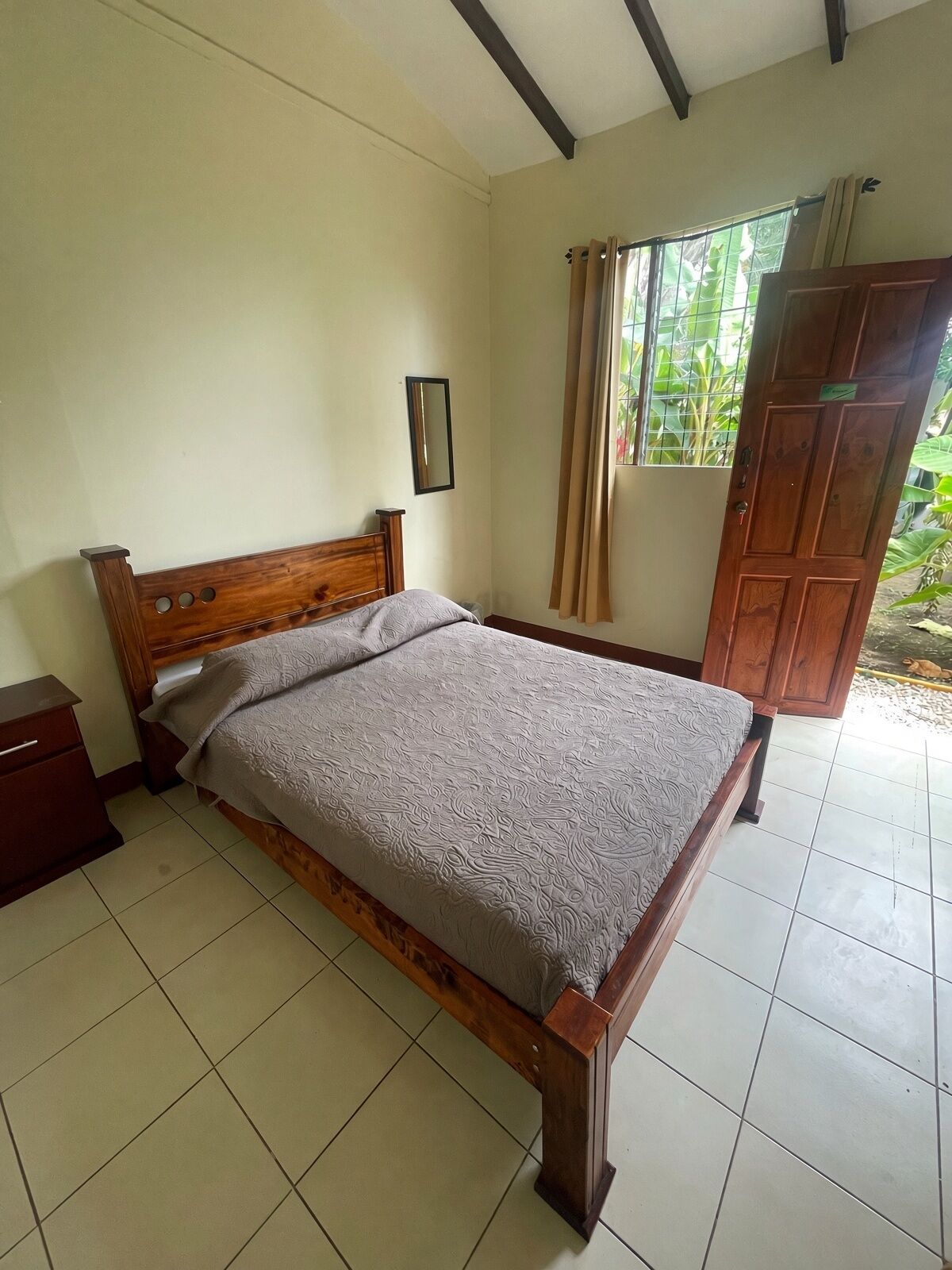 A single bed with a wooden frame and a patterned bedspread in a room with tiled flooring and a window with curtains.